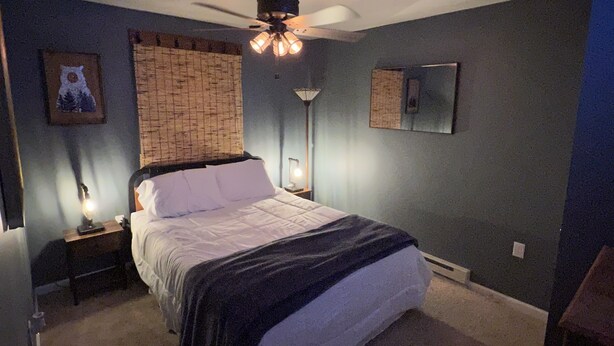 Un dormitorio con cama, ventilador de techo, mesita de noche, lámpara y un cuadro colgado en la pared.