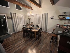 A dining area with a wooden table and chairs, a chandelier, and a kitchen area with a counter and stools.