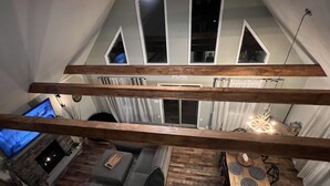Un espacio habitable estilo loft con techo alto, vigas de madera a la vista, una zona de comedor con mesa y sillas de madera, un sofá gris y un televisor al fondo.