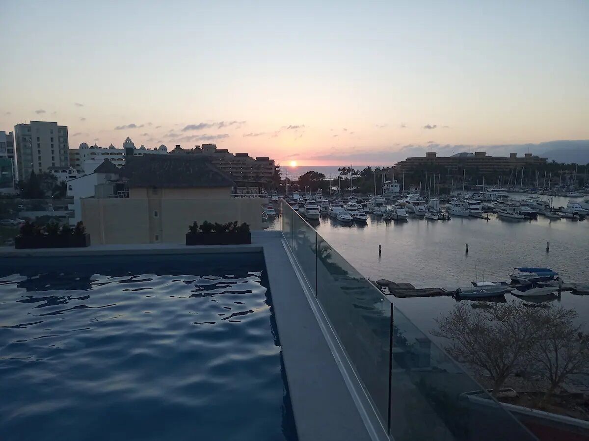 A marina with boats docked, a sunset in the background, and buildings in the distance.