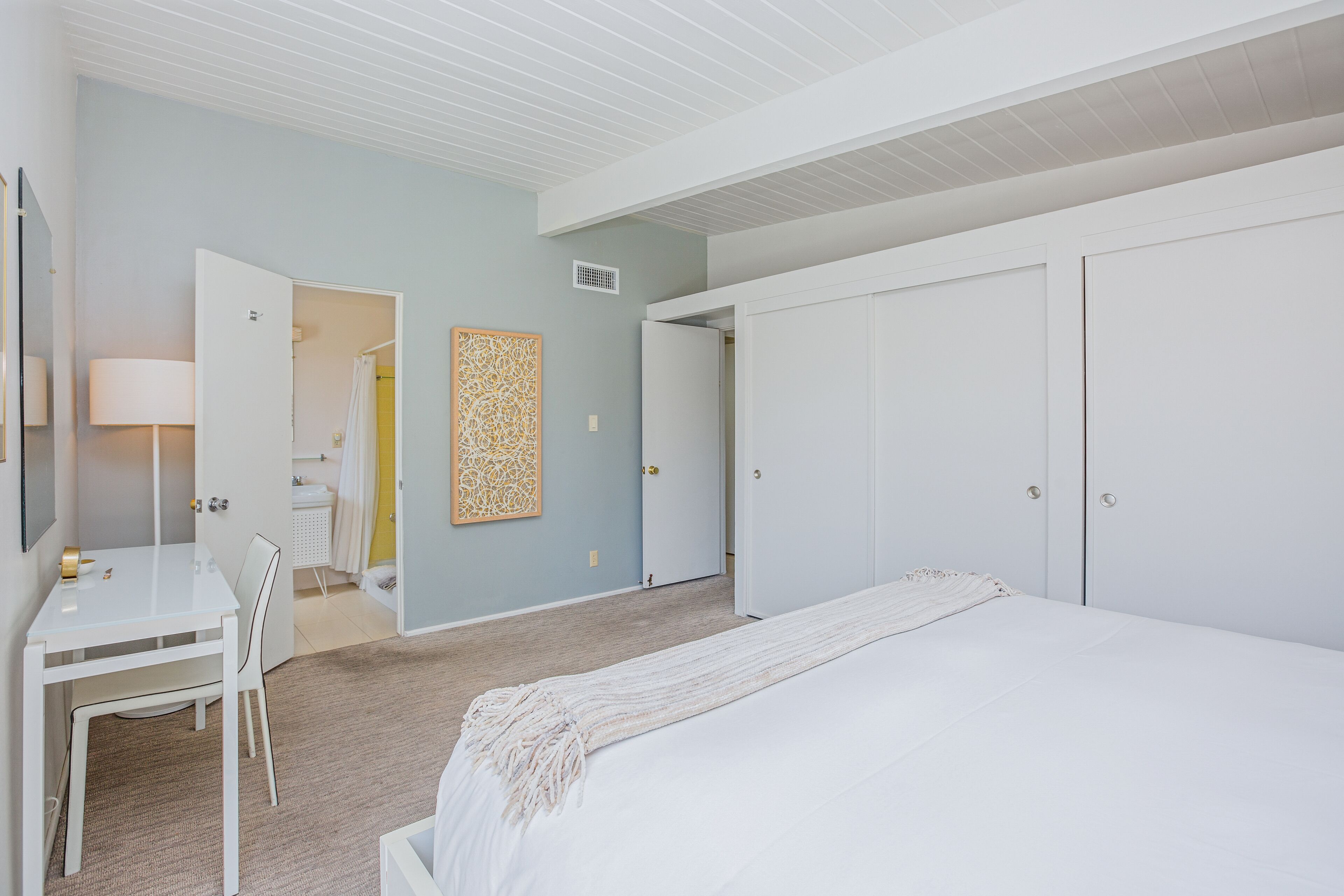 A bedroom with a bed, a desk, and a chair. There is a bathroom visible through an open door.