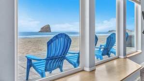 A room with a large window overlooking a beach with blue chairs and a rock formation.