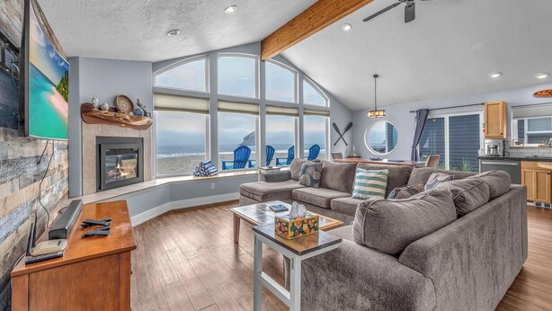 A spacious living room with a large window overlooking the beach, a fireplace, and a modern kitchen in the background.