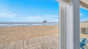 A beach view from a covered area with a wooden deck, a clear sky, and a rock formation in the distance.