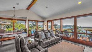 A spacious living room with a leather sofa, a wooden coffee table, and a view of a lake and mountains through large windows. - Incredible Ocean/Esturary Views, Large Covered Deck, Community Pool and Hot Tub (Pacific City)