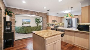 A modern kitchen with a central island, wooden cabinets, and a green patterned sofa in the background.