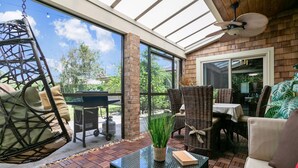 A sunlit patio with wicker furniture, a hanging chair, and a glass table with a plant.