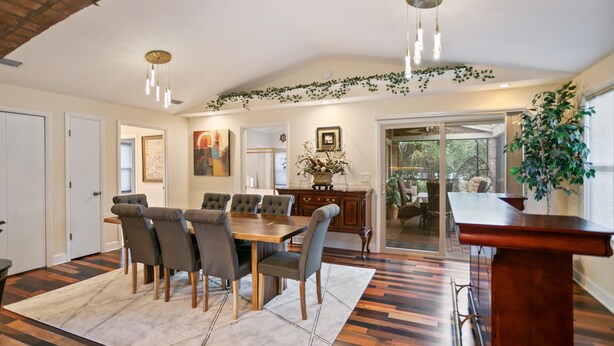 A dining area with a wooden table and chairs, a kitchen in the background, and a view through a glass door.
