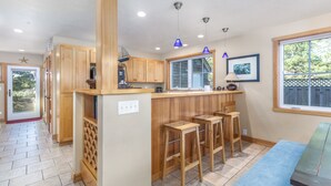 A kitchen with wooden cabinets, a central island with bar stools, a window with a view of greenery, and a door leading outside. - Delightfully Decorated Home Walking Distance to the Beach, Fireplace, Deck, BBQ (Pacific City)