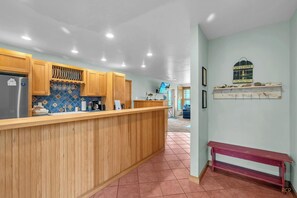 A kitchen with wooden cabinets, a countertop, and a tiled backsplash. There is a bench and a shelf with decorative items.