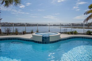 A swimming pool with a hot tub, surrounded by a fence, and a view of a body of water and buildings in the background. - Sea of Dreams. Remodeled Lakefront 4 Bed, Heated Pool, Golf Cart and Bikes! (Destin)