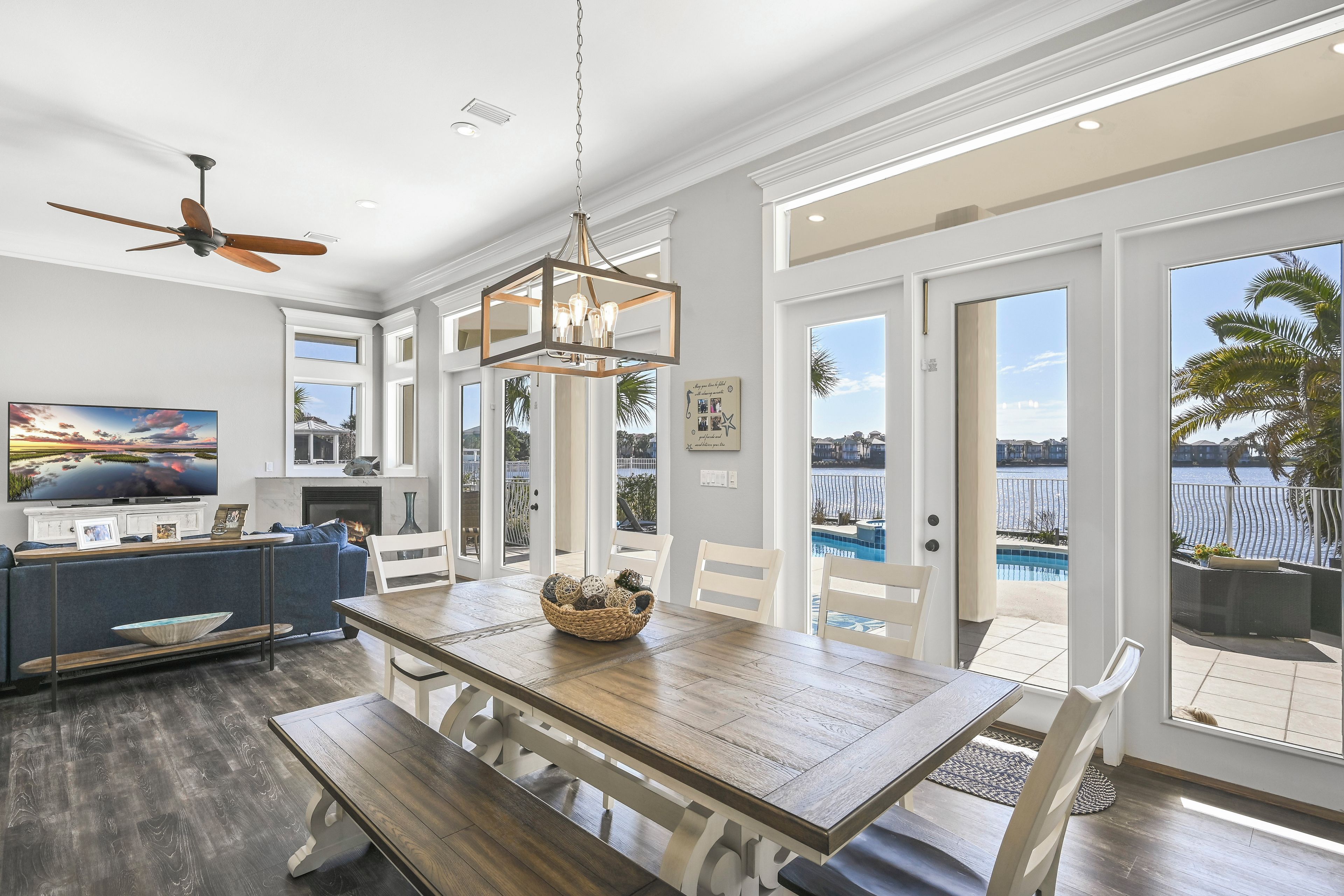 A spacious living area with a wooden dining table, white chairs, a ceiling fan, and a large window overlooking a pool.