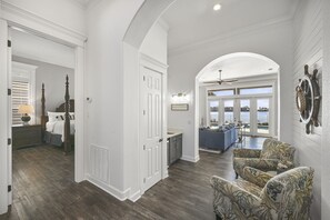 A spacious living area with a large window overlooking a pool, a cozy seating area with patterned chairs, and a kitchen with a sink and countertop. - Sea of Dreams. Remodeled Lakefront 4 Bed, Heated Pool, Golf Cart and Bikes! (Destin)