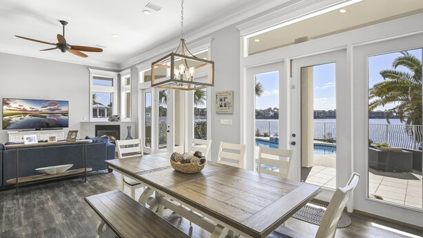 A spacious living area with a wooden dining table, white chairs, a ceiling fan, and a large window overlooking a pool.