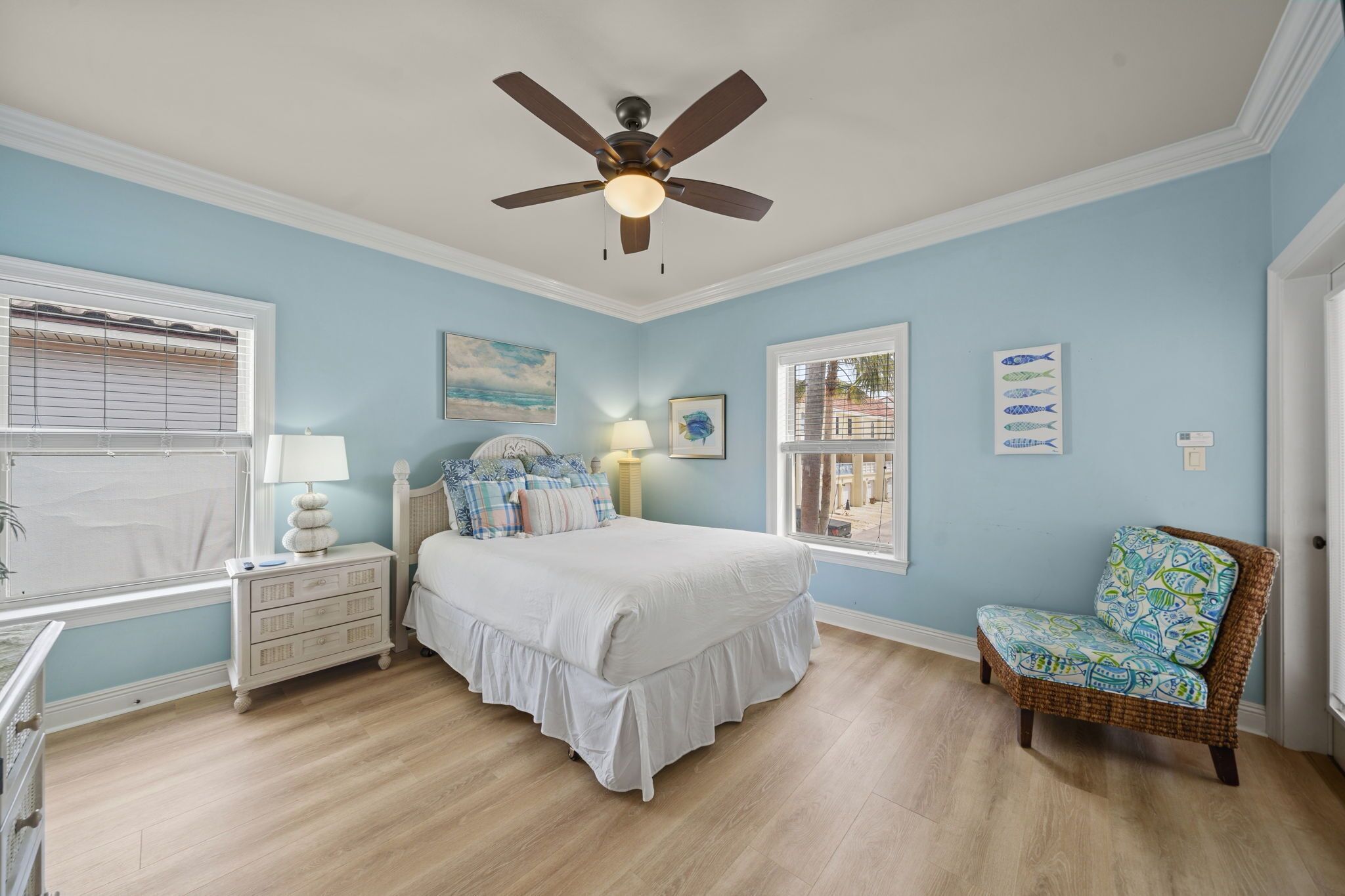 Une chambre à coucher comprenant un lit, des tables de chevet, une chaise, un ventilateur de plafond et des œuvres d’art accrochées aux murs.
