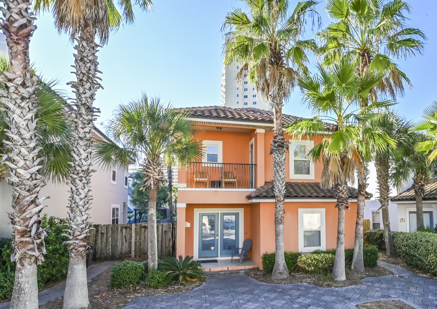 A two-story building with a balcony, surrounded by palm trees and a wooden fence.