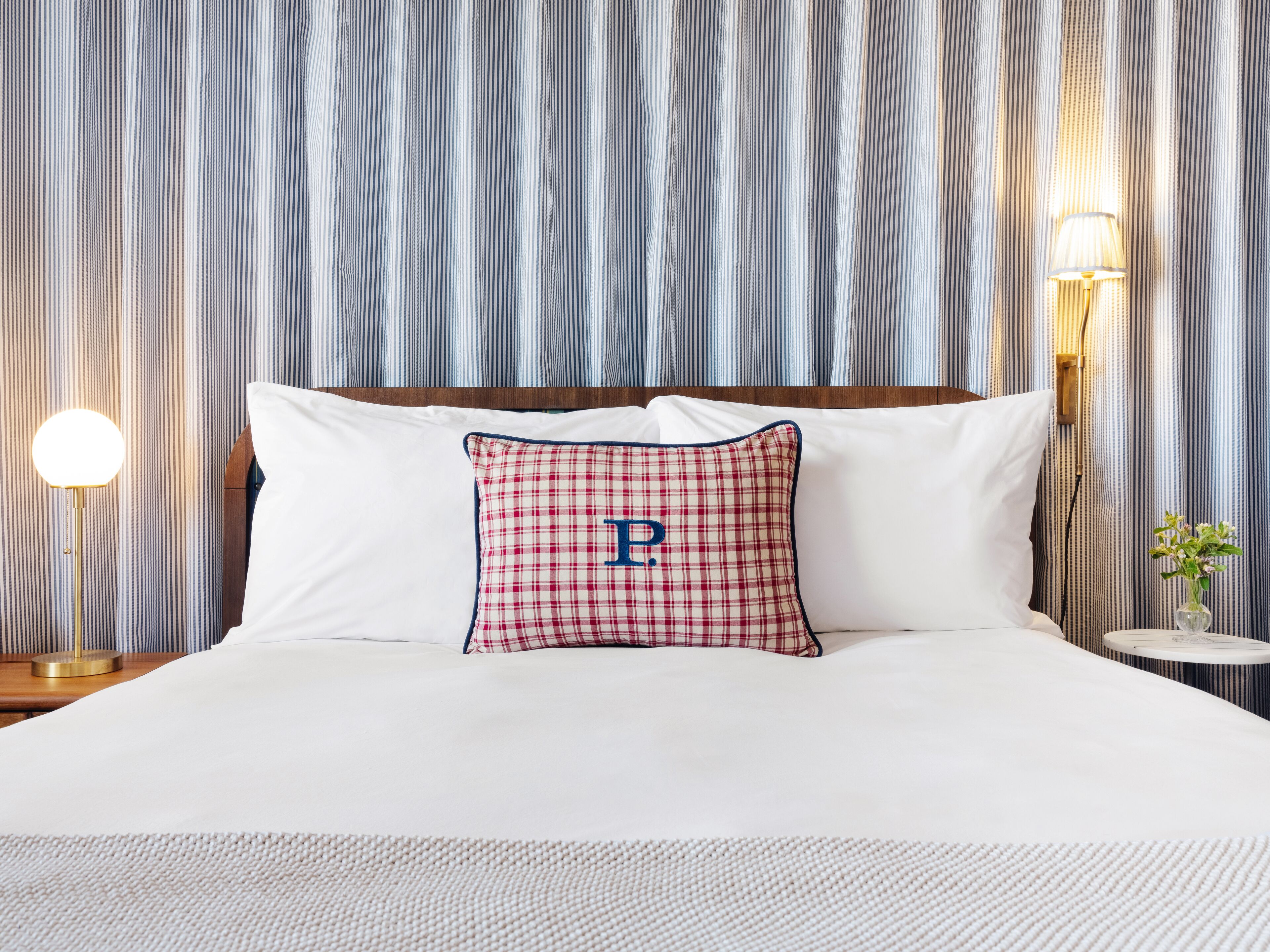 Un lit bien fait, avec du linge de lit blanc, un coussin à carreaux orné de la lettre « P » et un papier peint à rayures.