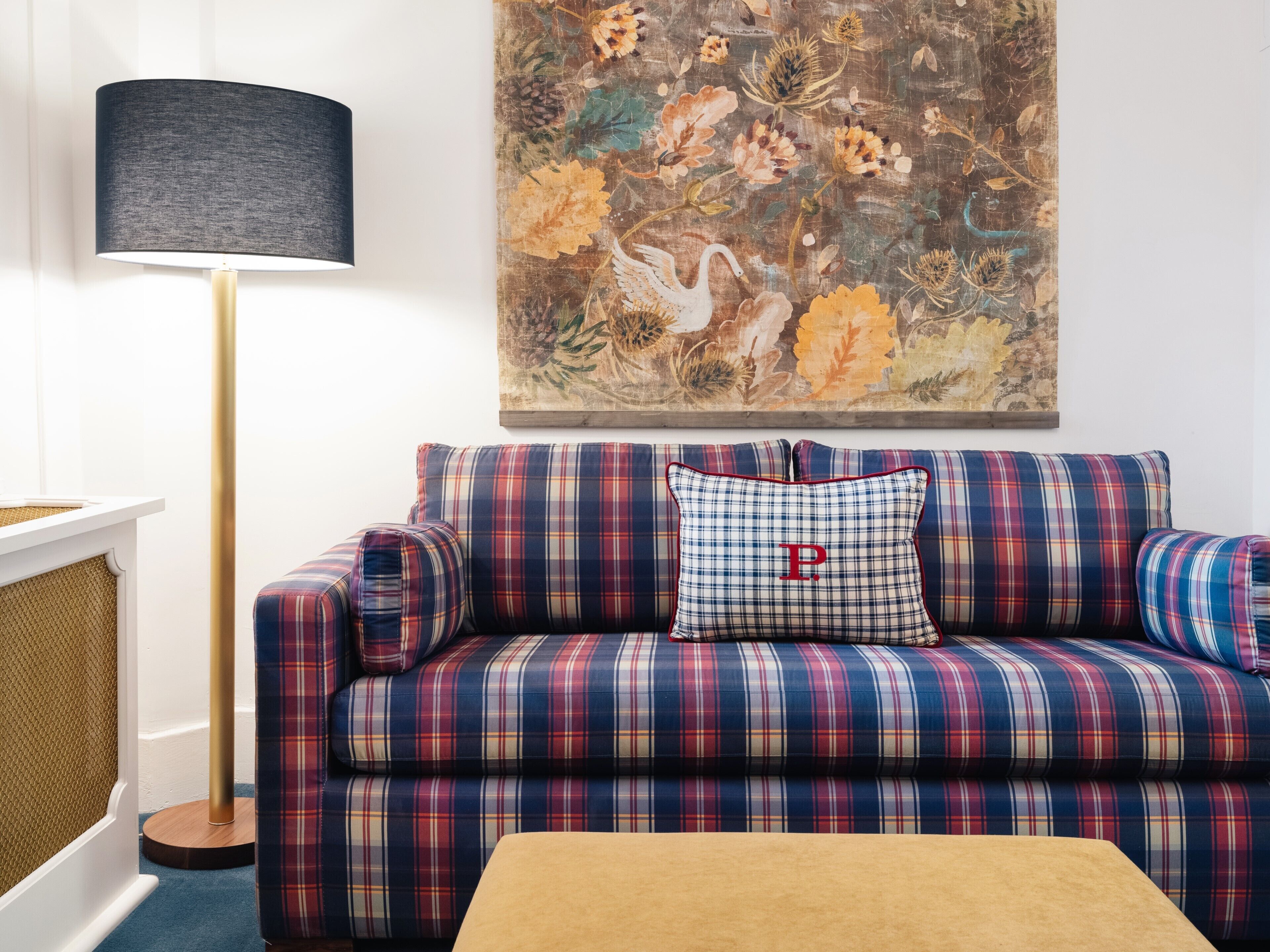 A plaid-patterned sofa with a cushion featuring a large 'P' logo, a floor lamp with a dark shade, and a floral painting on the wall.