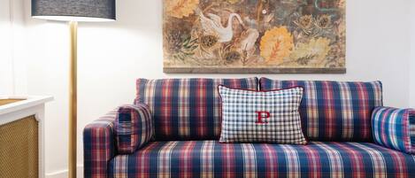 A plaid-patterned sofa with a cushion featuring a large 'P' logo, a floor lamp with a dark shade, and a floral painting on the wall.