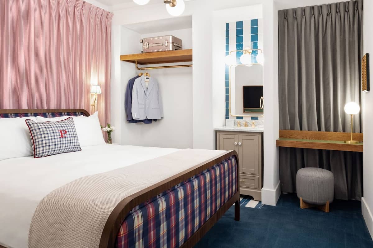 Ein Schlafzimmer mit einem Bett, einem Nachttisch, einem Spiegel, einer Kommode, einem kleinen Hocker und einem Fenster mit Vorhängen.