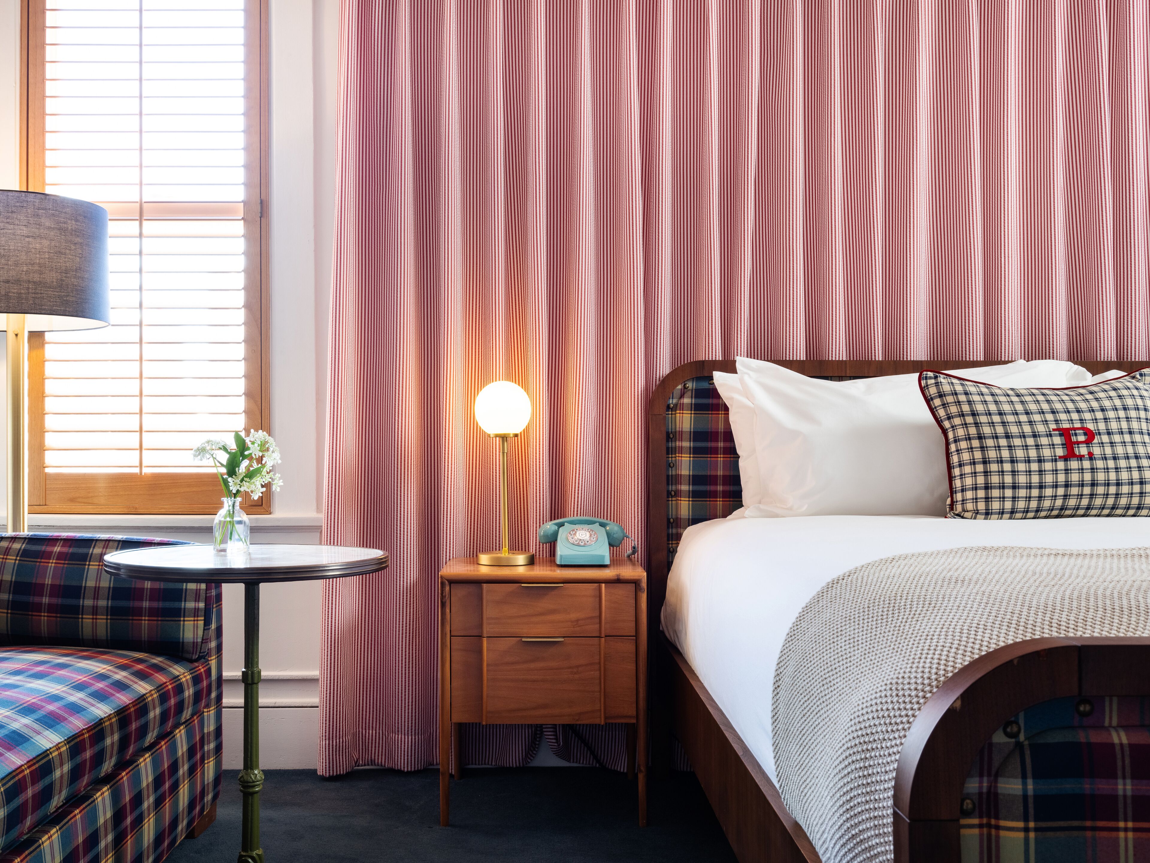 A hotel room with a plaid sofa, a bedside table with a lamp, a vintage telephone, and a bed with a plaid blanket and pillows.