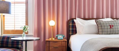 A hotel room with a plaid sofa, a bedside table with a lamp, a vintage telephone, and a bed with a plaid blanket and pillows.