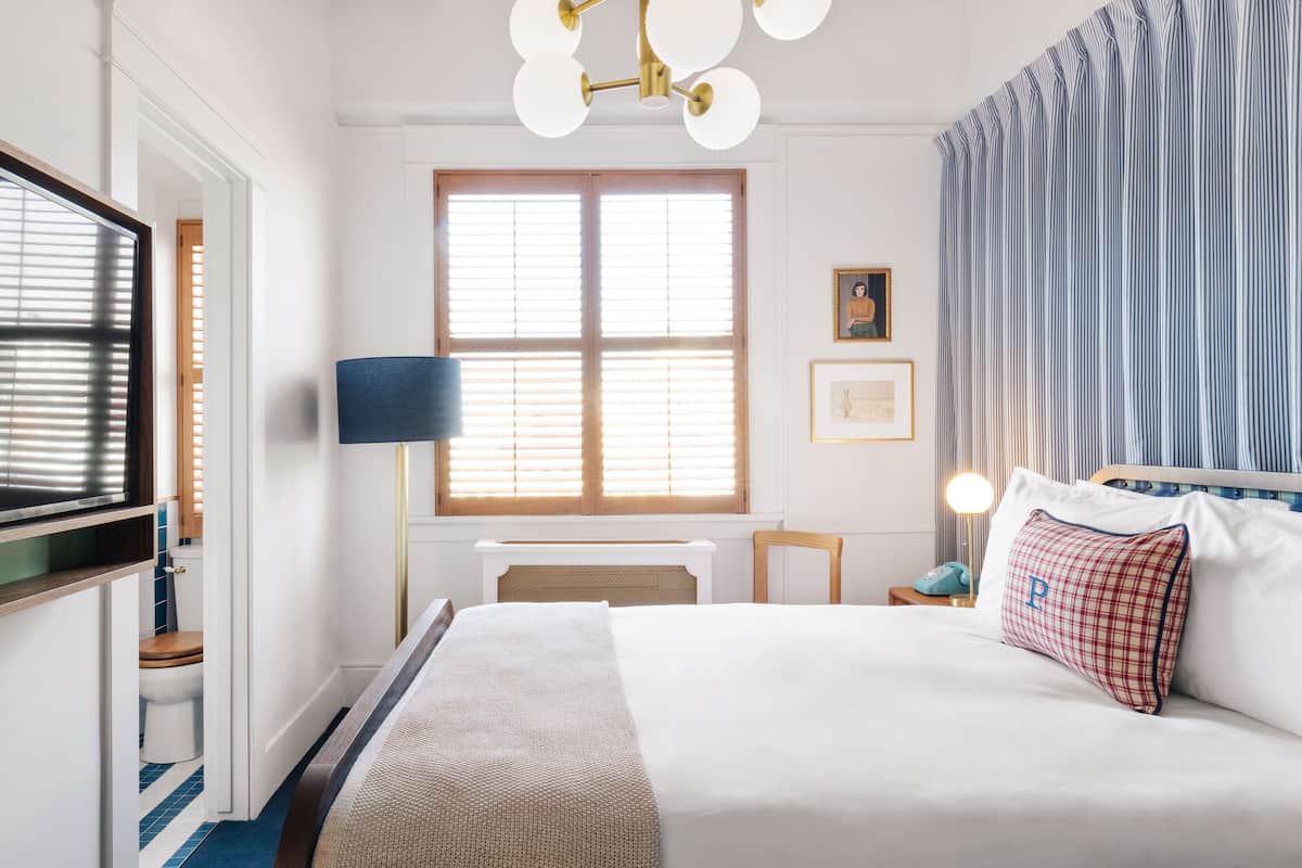 Ein Hotelzimmer mit einem großen Bett, einem Schreibtisch, einem Stuhl, einer Lampe und einem Fenster mit Jalousien.