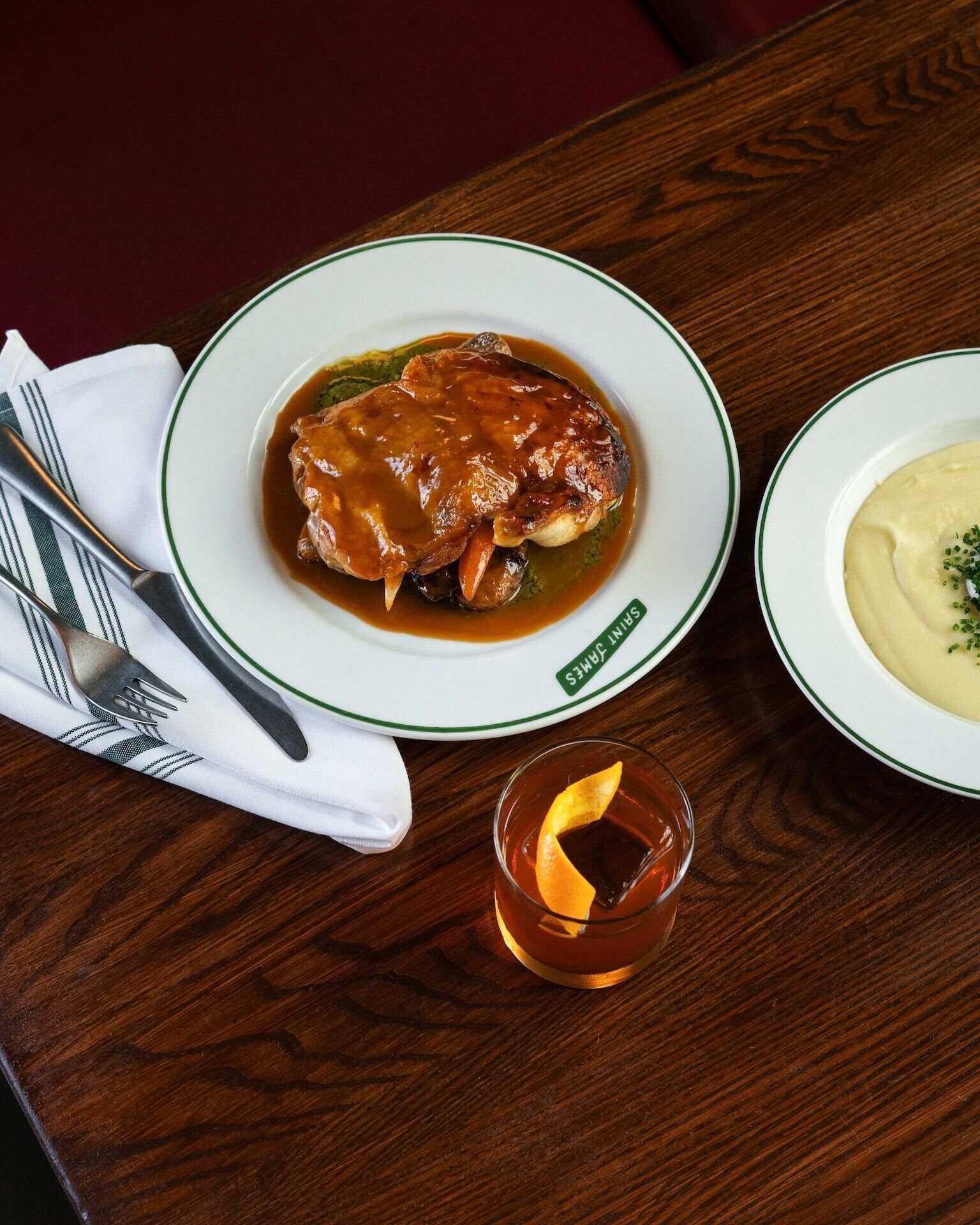 A plate of food with gravy, a glass of drink, and a napkin with cutlery on a wooden table.