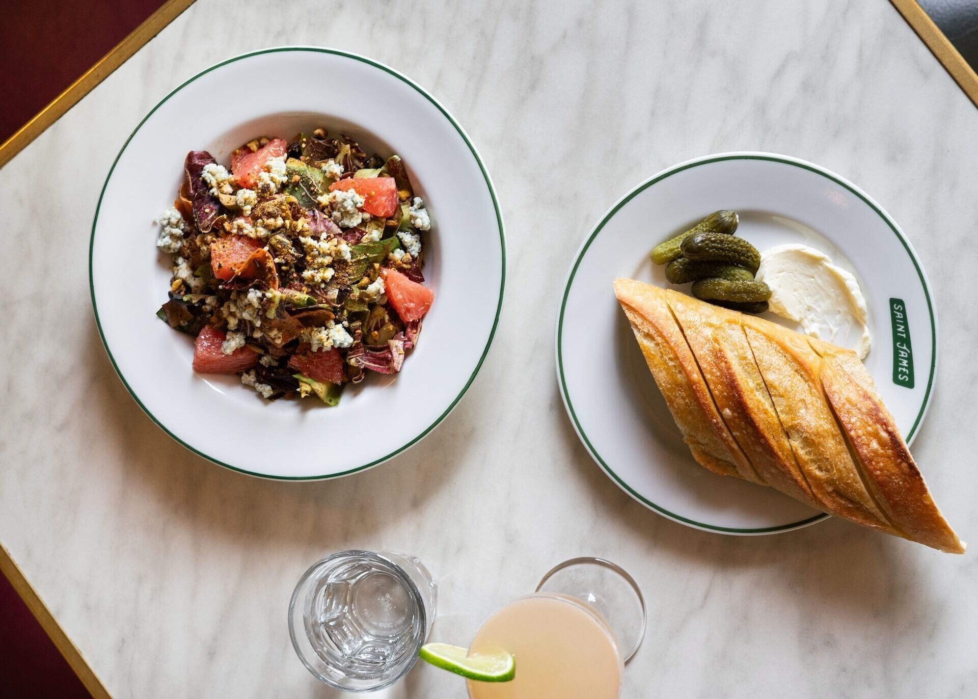 Two plates of food on a marble table, one with a salad and the other with bread and pickles.