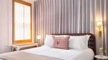 A neatly made bed with a plaid pillow, a bedside lamp, and a window with blinds.