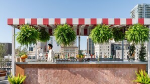 A rooftop bar with a bartender, potted plants, and a city view.