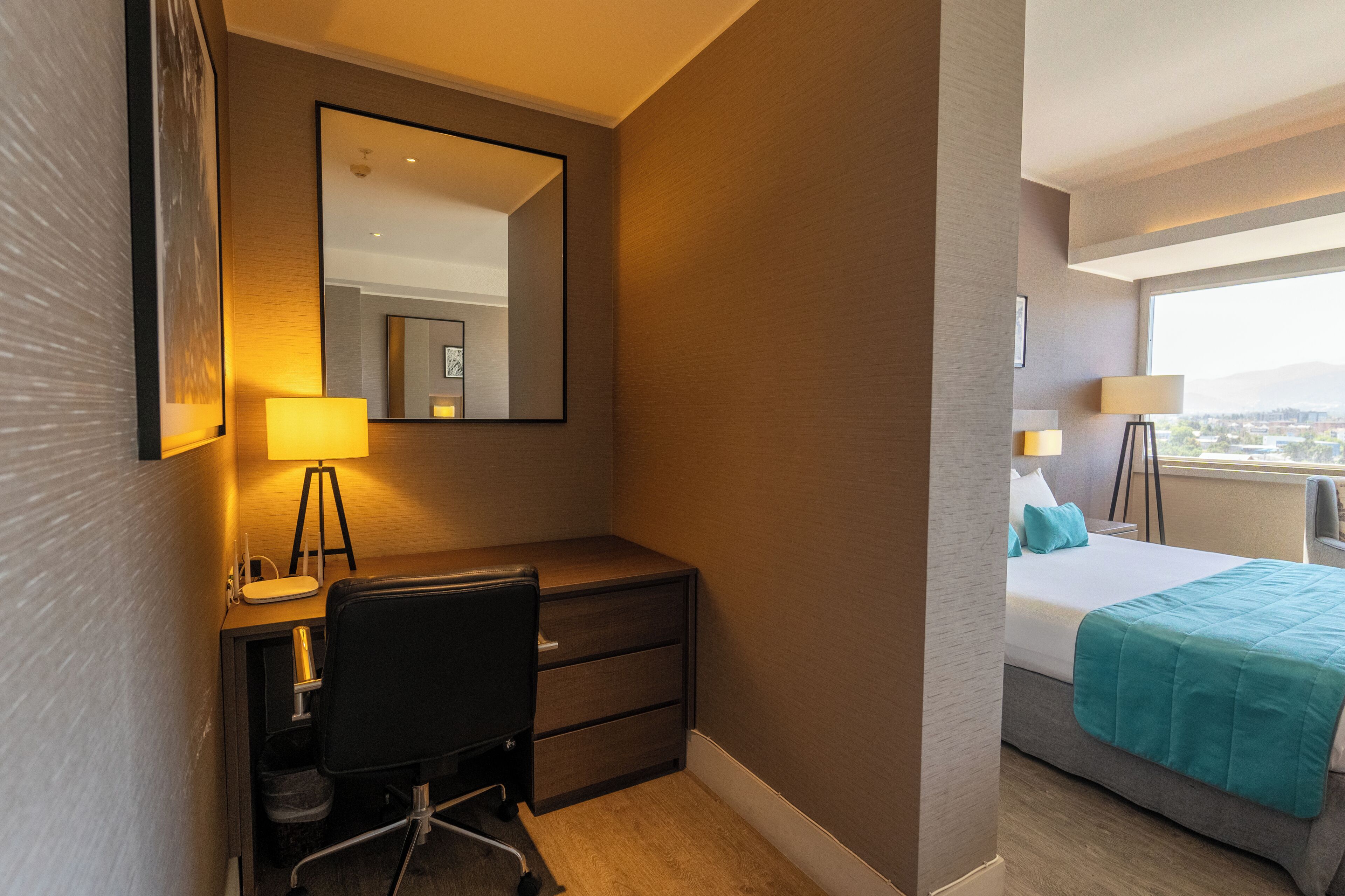 Una habitación de hotel con escritorio, silla, cama, lámparas de noche y un ventanal grande.