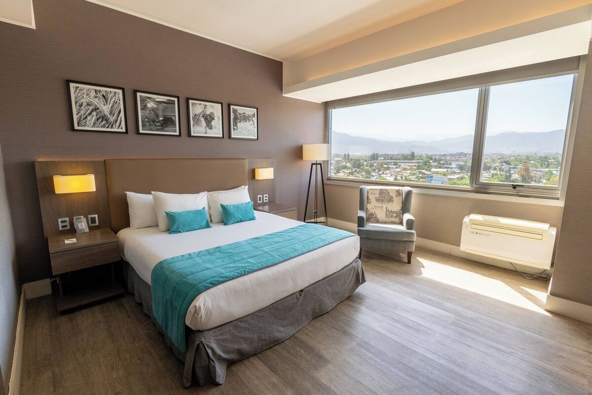 Habitación de hotel con una cama grande, lámparas de noche, una silla y vistas a un paisaje urbano a través de un ventanal amplio.