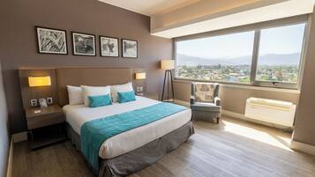 Une chambre d’hôtel avec un grand lit, des lampes de chevet, un fauteuil et une vue sur le paysage urbain, visible à travers une grande fenêtre.