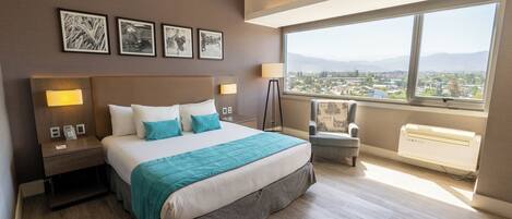 A hotel room with a large bed, bedside lamps, a chair, and a view of a cityscape through a large window.