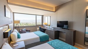 Habitación de hotel con dos camas, un escritorio, un televisor y un ventanal con vista a la ciudad.