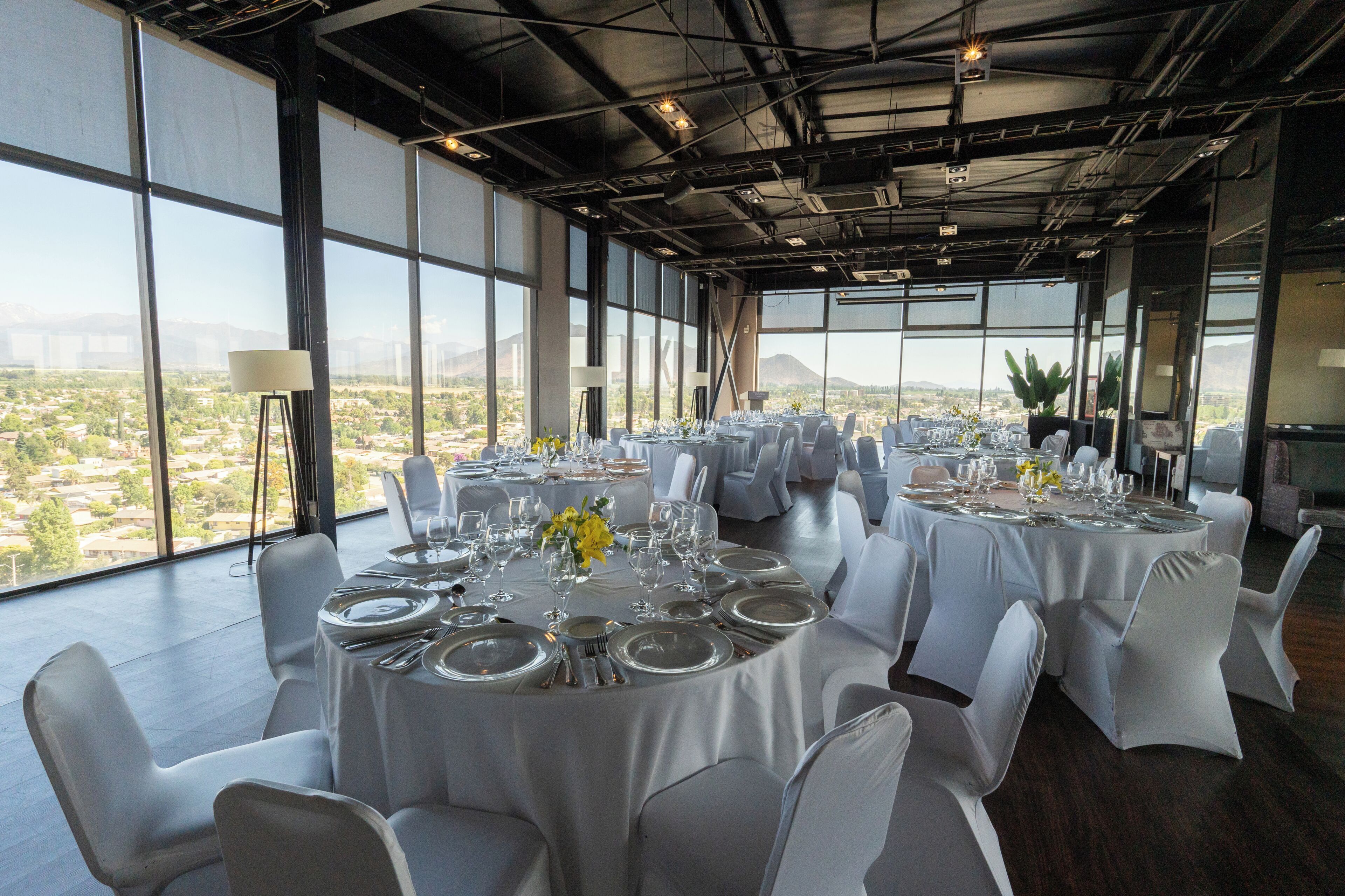 A spacious dining area with round tables set for a meal, large windows offering a view of a cityscape, and a high ceiling with exposed beams.