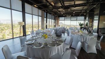 A spacious dining area with round tables set for a meal, large windows offering a view of a cityscape, and a high ceiling with exposed beams.