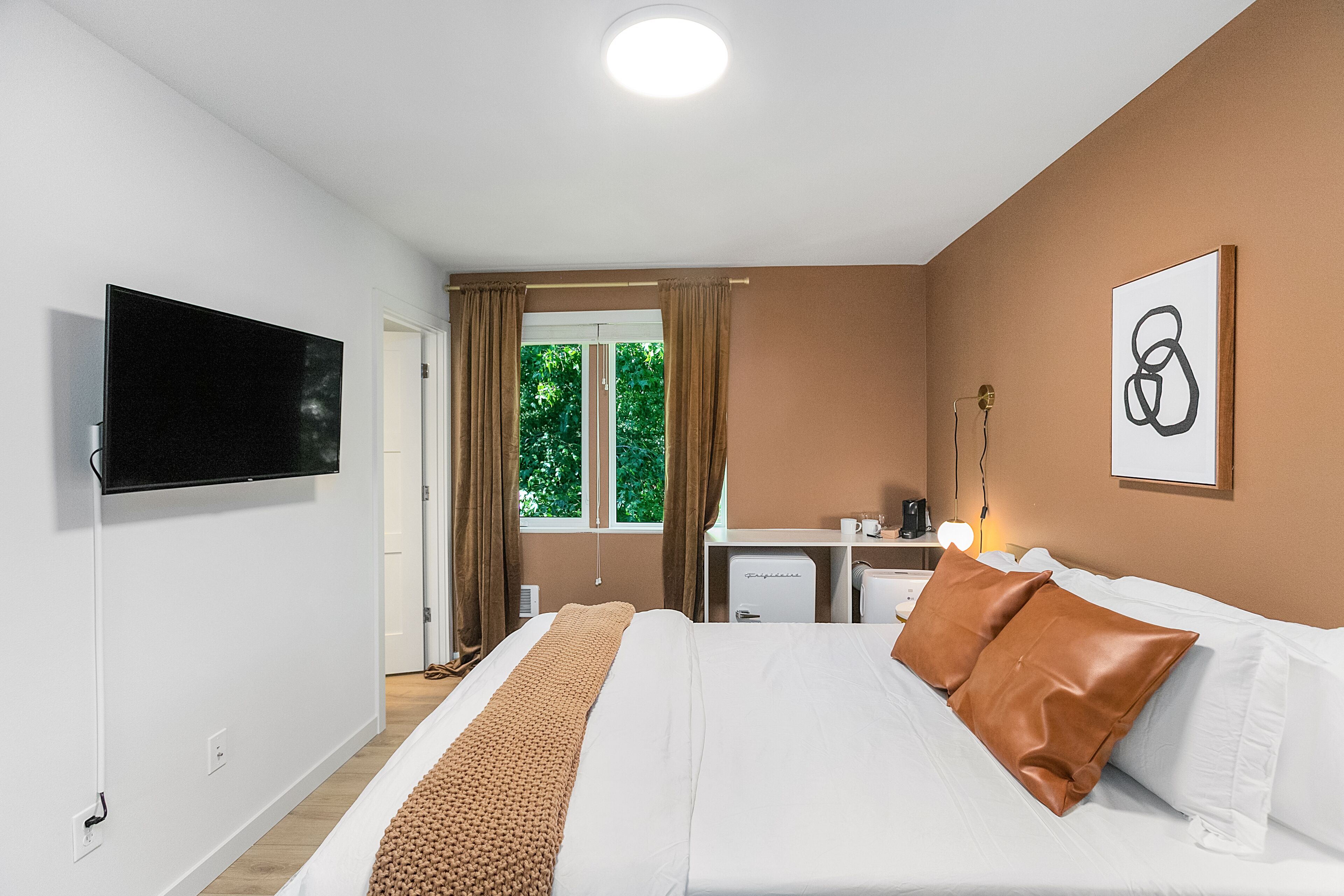 A hotel room with a bed, a flat-screen TV, a desk with a lamp, and a window with curtains.