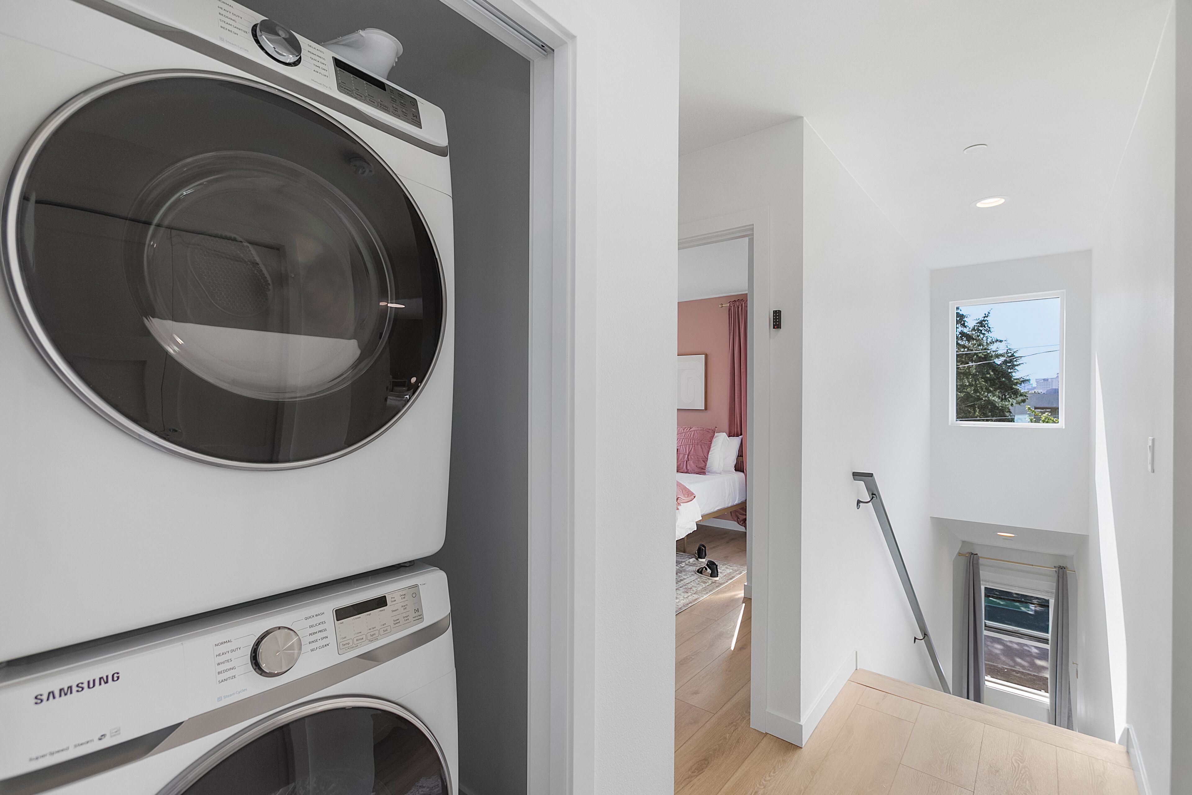 Een moderne wasruimte met een Samsung wasmachine en droger, een trap die leidt naar een slaapkamer met een bed en een raam, en een klein balkon met uitzicht.