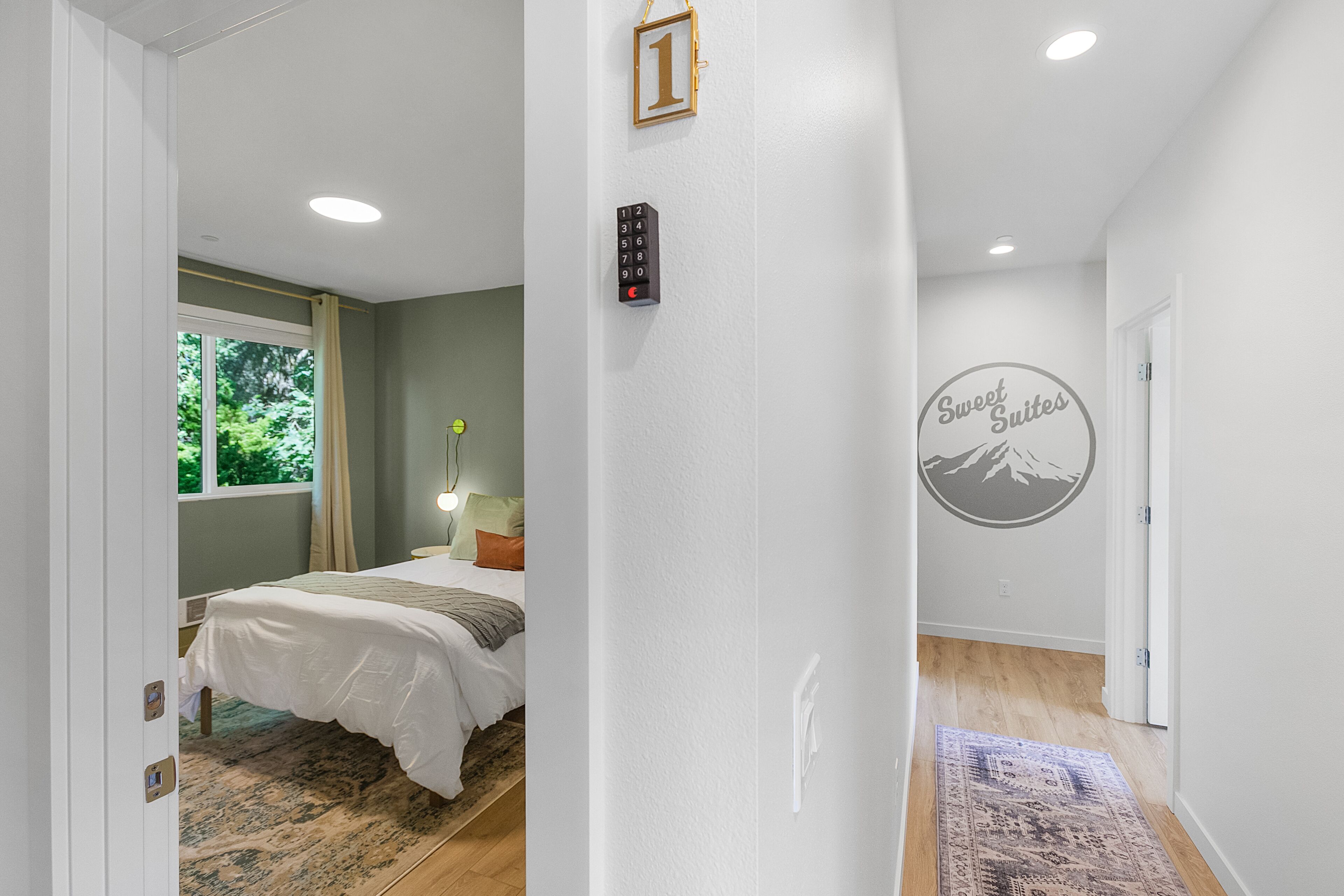 A hotel room with a bed, a bedside table with a lamp, a window with curtains, a wall-mounted phone, and a door with a sign.