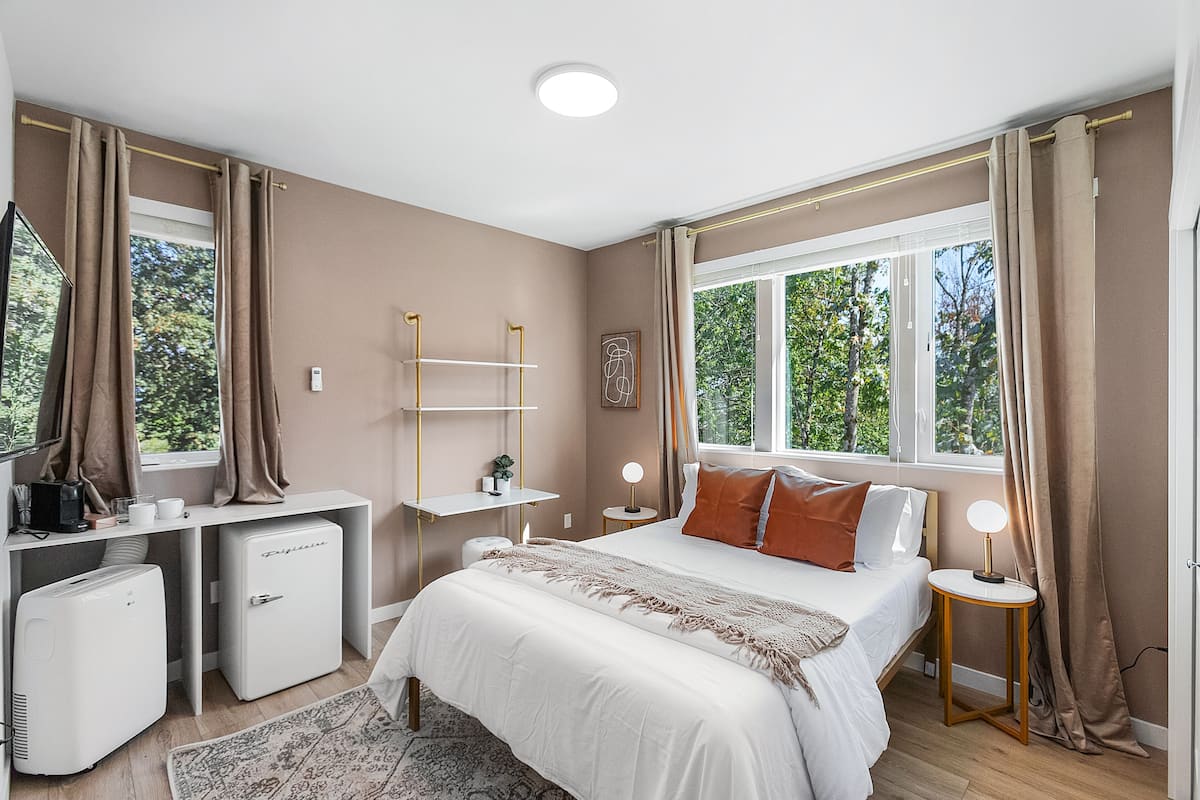 A bedroom with a bed, bedside tables, a desk, a refrigerator, and a window with curtains.