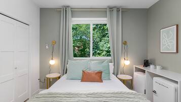 Un dormitorio con cama, mesitas de noche, escritorio, refrigerador y ventana con cortinas.