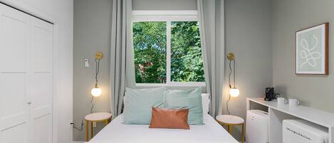 A bedroom with a bed, bedside tables, a desk, a refrigerator, and a window with curtains.