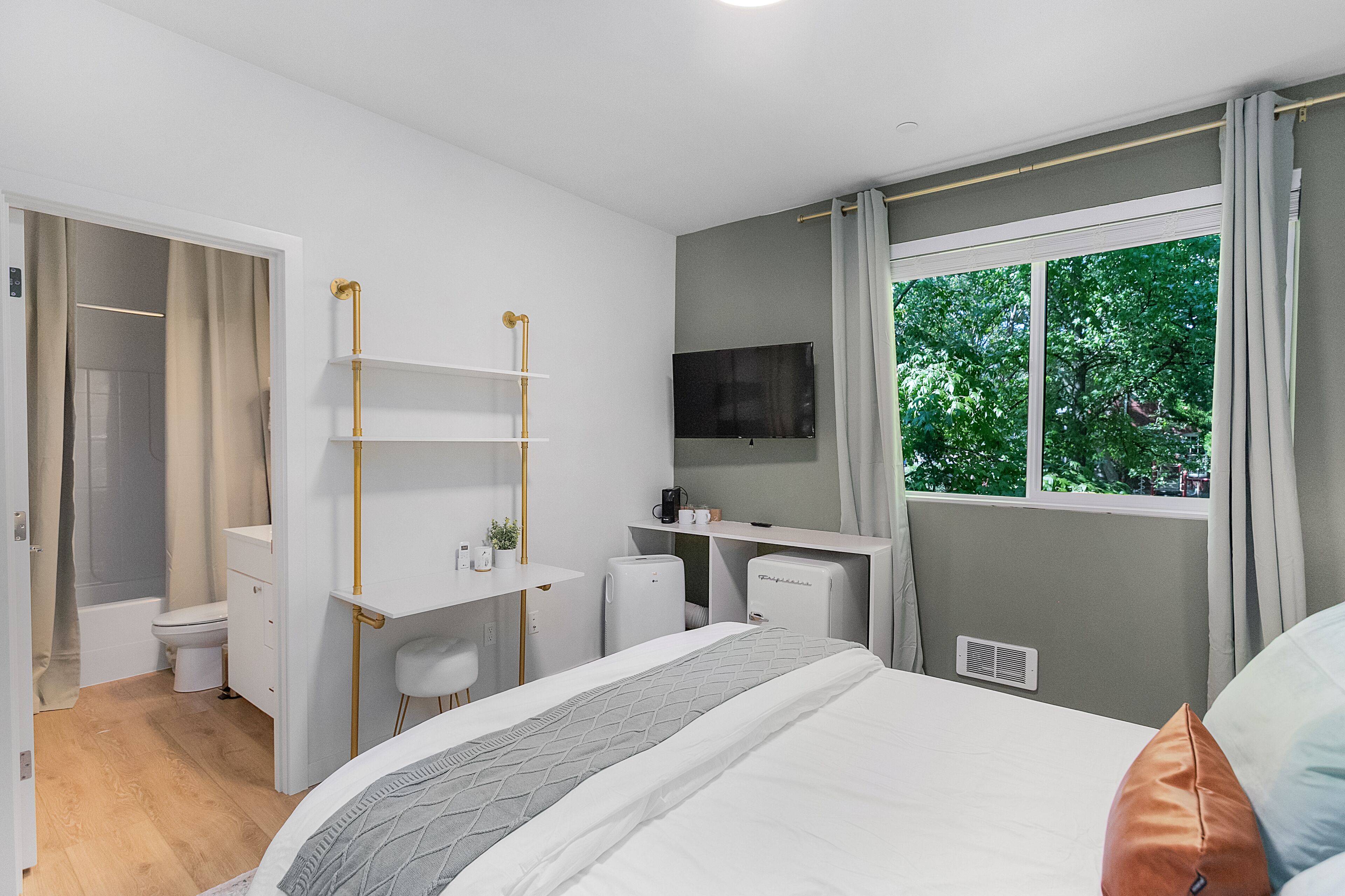Un dormitorio moderno con cama, escritorio, silla, televisor y ventana con vista a árboles.
