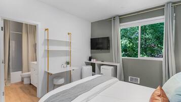 Un dormitorio moderno con cama, escritorio, silla, televisor y ventana con vista a árboles.