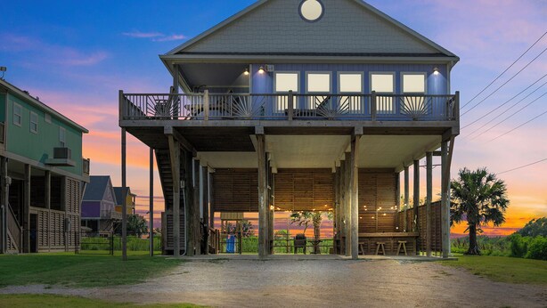 A two-story wooden building with a balcony, a covered outdoor seating area, and a view of the sunset. - Summer Special! Views! Beachside, HotTub, Arcade, FirePit, Pets, Family Retreat (Galveston)