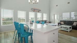 A spacious living area with a large white island, teal chairs, a black sofa, and a ceiling light fixture. - Summer Special! Views! Beachside, HotTub, Arcade, FirePit, Pets, Family Retreat (Galveston)