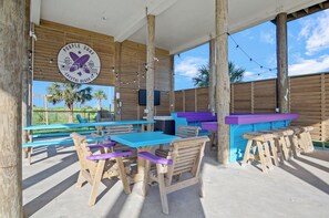 Zona de pati a l'aire lliure amb taules i cadires de fusta, un taulell de bar morat i un cartell que diu "Purple Surf Crystal Beaches".