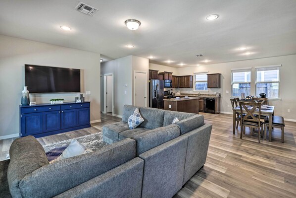 A spacious living room with a sectional sofa, a flat-screen TV, and a dining area with a table and chairs. - Family Home w/ Fenced Yard in Austin Suburbs! (Buda)